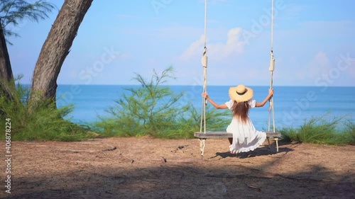 Dressed in a classical summer sundress, a woman with her back to the camera gently swings on a large wooden swing looking out at the ocean horizon. photo