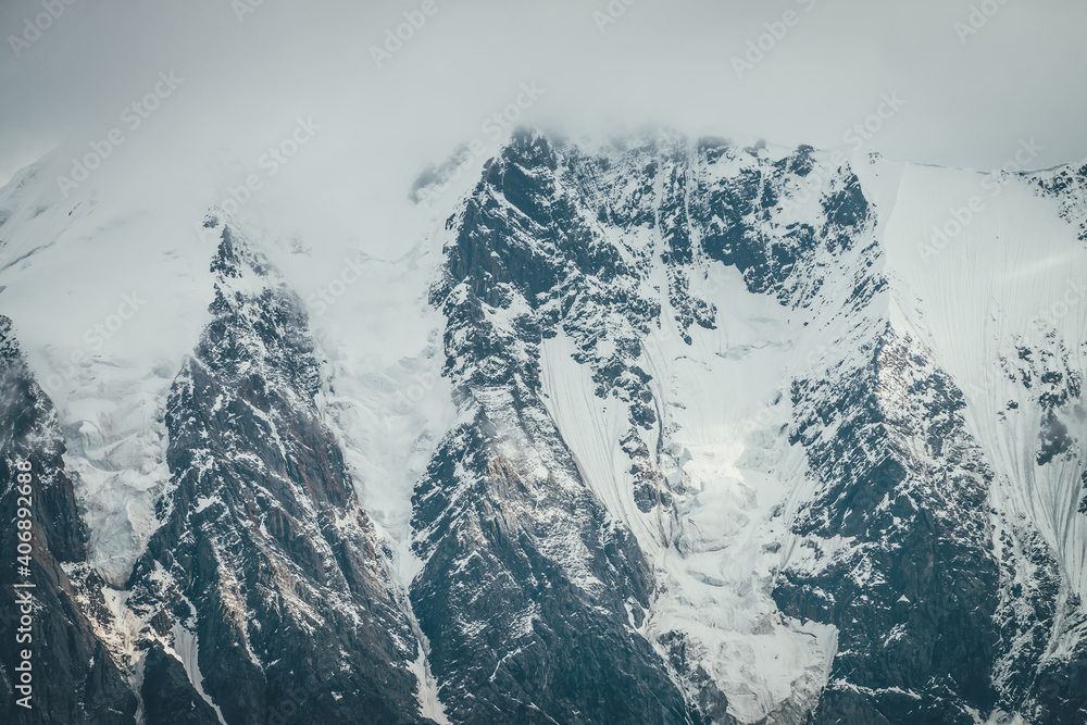 Obraz premium Atmospheric mountains landscape with big snowy mountain top with snow cornices in low clouds. Awesome minimal scenery with snow-white high pinnacle in overcast weather. Giant mountain wall in clouds.