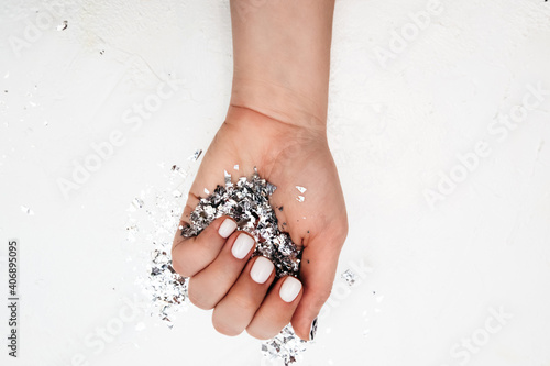 light-skinned hand with perfect manicure with milky color gel polish holds large handful of silver foil sequins on white background, selective focus
