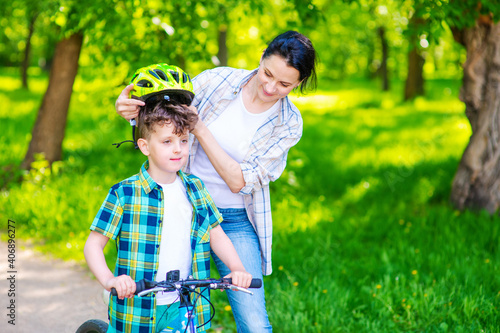 Mom helping son to wear a bicycle helmet in a summer park. Weekend Leisure Concept