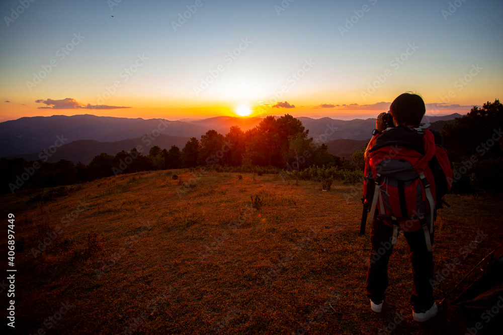 A man who loves adventure and photography. Stand on the top of the mountain with a tent, enjoy the beautiful view at sunset.