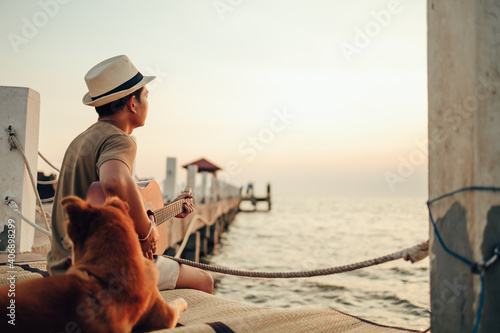 A man wear straw hat and playing guitar music song near the sea sunset with a dog pet.