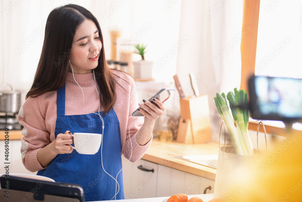 Young Asian woman vlogger use smart phone and hold coffee cup, recording video for food channel in her kitchen, online cooking class at home, video blogging concept