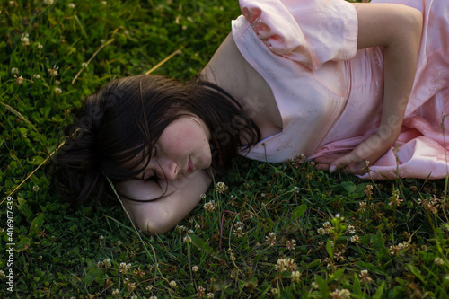 Portret of a girl with dark hair in a pink dress lies and sleeps in the green grass with white clover flowers on a warm summer evening. Photo of beautiful sleeping woman in the fild with wildflowers.
