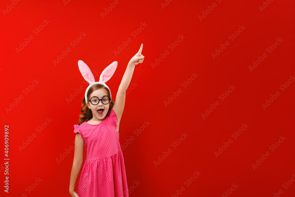 Happy cute little girl wearing easter bunny ears and glasses on red background. The child points to an empty copy space.