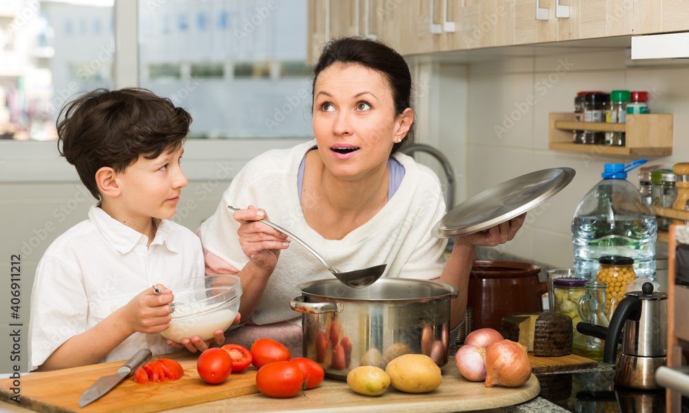 Positive woman and her preteen son preparing food and funny talking in cosy kitchen interior