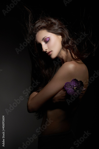 Beauty portrait of a nude woman with a purple flower in her hands on a dark background. Natural cosmetics, natural makeup