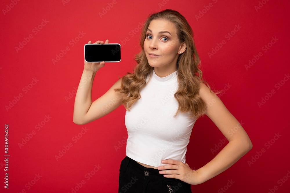 beautiful young blonde woman wearing white t-shirt isolated on red background with copy space holding smartphone showing phone in hand with empty screen for mockup looking at camera beautiful young blonde woman wearing white t-shirt isolated on red background with copy space holding smartphone showing phone in hand with empty screen for mockup looking at camera