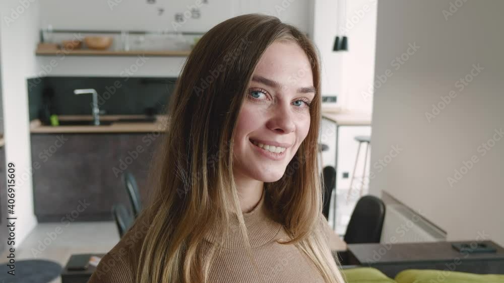 Portrait of attractive young woman model looking at camera and smiling at home. Portrait if beautiful female with pretty face and blue eyes stand alone indoors at living room. Steadicam shot. Portrait of attractive young woman model looking at camera and smiling at home. Portrait if beautiful female with pretty face and blue eyes stand alone indoors at living room. Steadicam shot.
