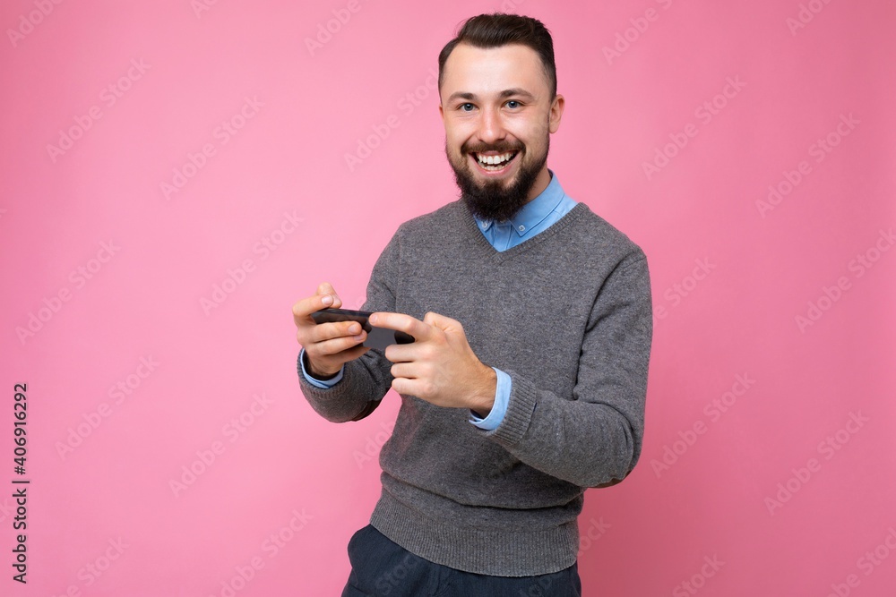 Positive smiling handsome good looking brunet bearded young man wearing grey sweater and blue shirt isolated on pink background with empty space holding in hand and using mobile phone communicating Positive smiling handsome good looking brunet bearded young man wearing grey sweater and blue shirt isolated on pink background with empty space holding in hand and using mobile phone communicating