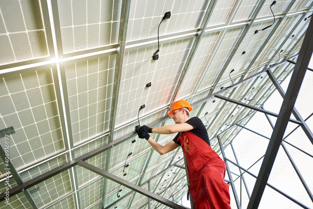 Fototapeta premium Solar power station with large volumes. The young man inside her is setting up a system. A guy in an orange uniform and a helmet on his head. Solar station development concept.