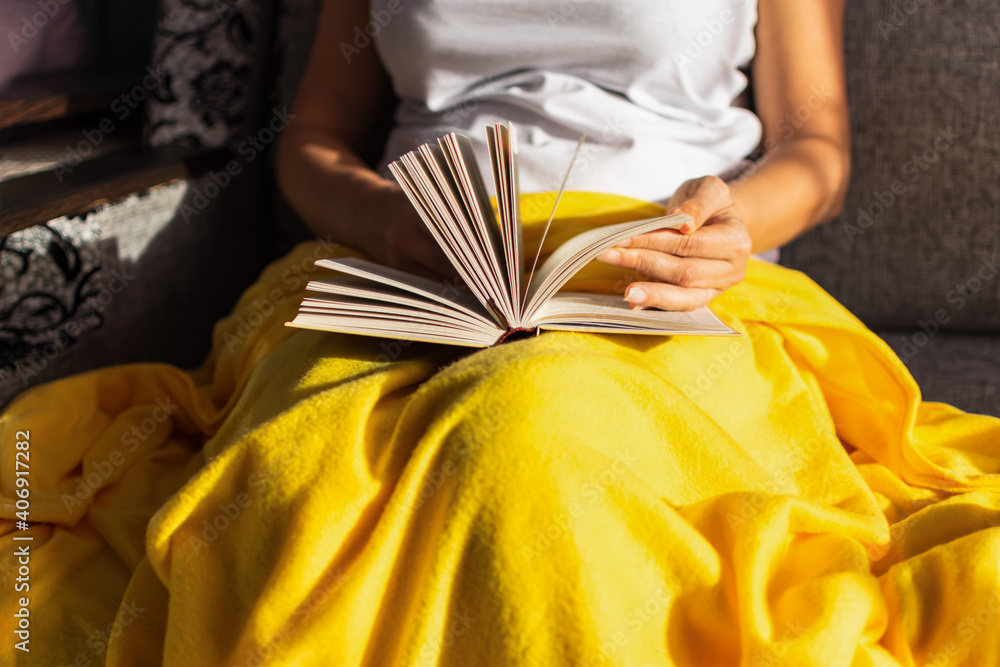 A girl in a white T-shirt is sitting on the couch and reading a book, covered with a beautiful yellow blanket. Bright sunlight, hard shadows.The concept of comfort, happiness.Trendy illuminating color