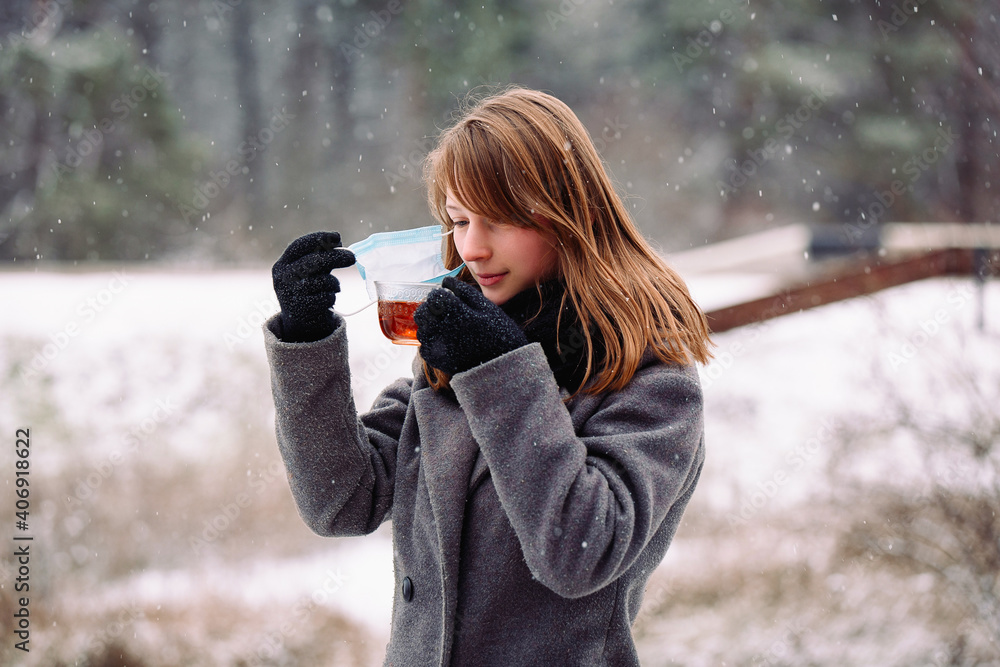 Caucasian girl removed the medical mask from her face to drink hot tea and keep warm outdoor in the winter cold forest. Caucasian girl removed the medical mask from her face to drink hot tea and keep warm outdoor in the winter cold forest.