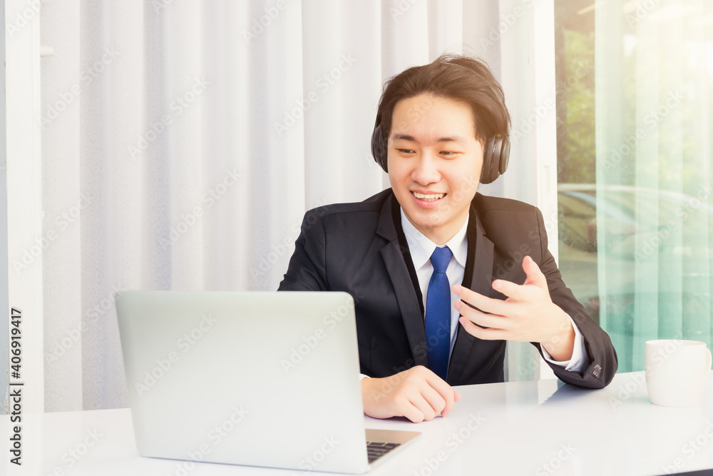 Work from home, Asian young businessman smile wearing headphones and suit video conference call or facetime on desk and raise his hand to explain to colleagues at home office Work from home, Asian young businessman smile wearing headphones and suit video conference call or facetime on desk and raise his hand to explain to colleagues at home office