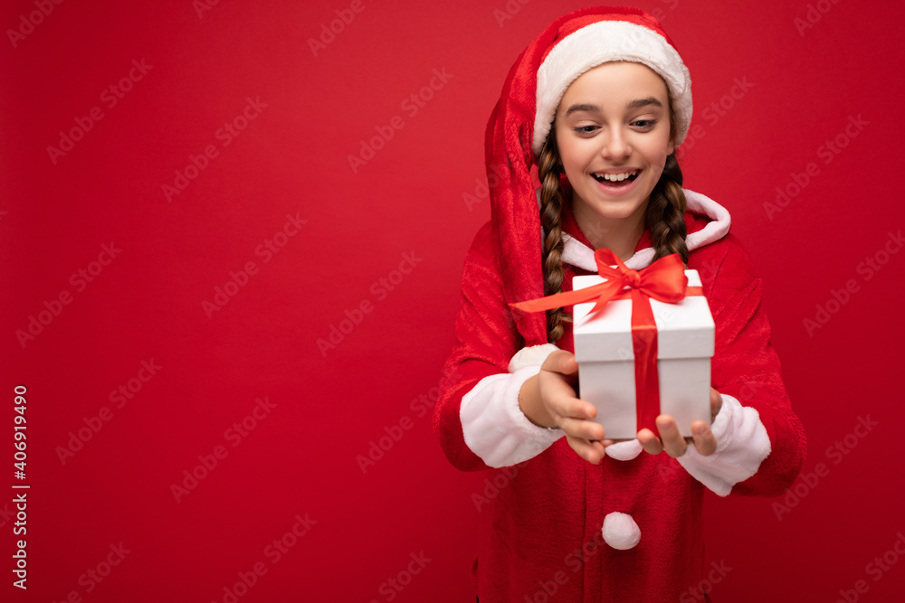 Shot of beautiful happy smiling brunette girl isolated over red background wall wearing new year santa claus outfit holding white gift box with red ribbon Shot of beautiful happy smiling brunette girl isolated over red background wall wearing new year santa claus outfit holding white gift box with red ribbon