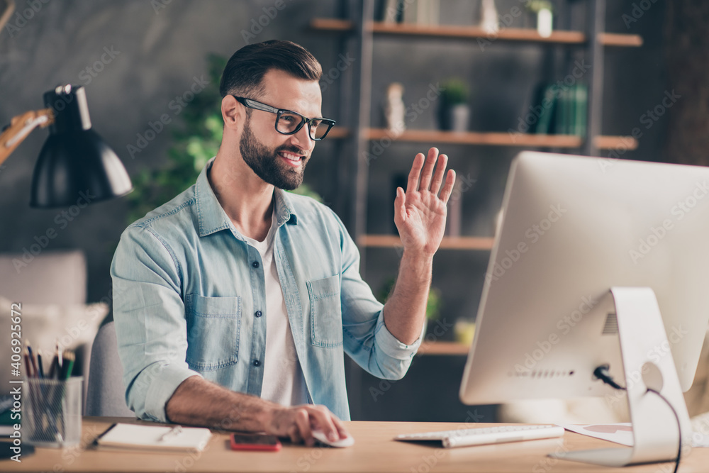 Photo portrait of guy at online meeting waving hand sitting at table in modern industrial office indoors Photo portrait of guy at online meeting waving hand sitting at table in modern industrial office indoors