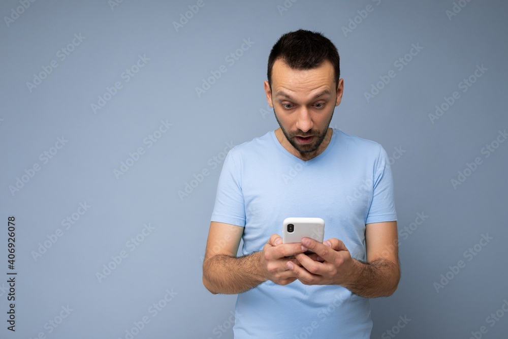 Photo of amazed handsome young man with beard wearing everyday blue t-shirt isolated over blue background holding and using mobile phone communication online on the internet looking at gadjet display