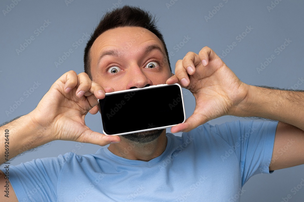 Closeup portrait of shocked handsome young unshaven brunet man with beard wearing everyday blue t-shirt isolated over blue background holding and showing mobile phone with empty display for mockup