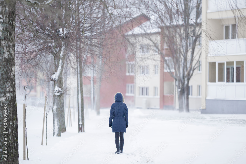 One young adult woman in dark warm coat walking on snow covered sidewalk in city. White winter day. Back view.