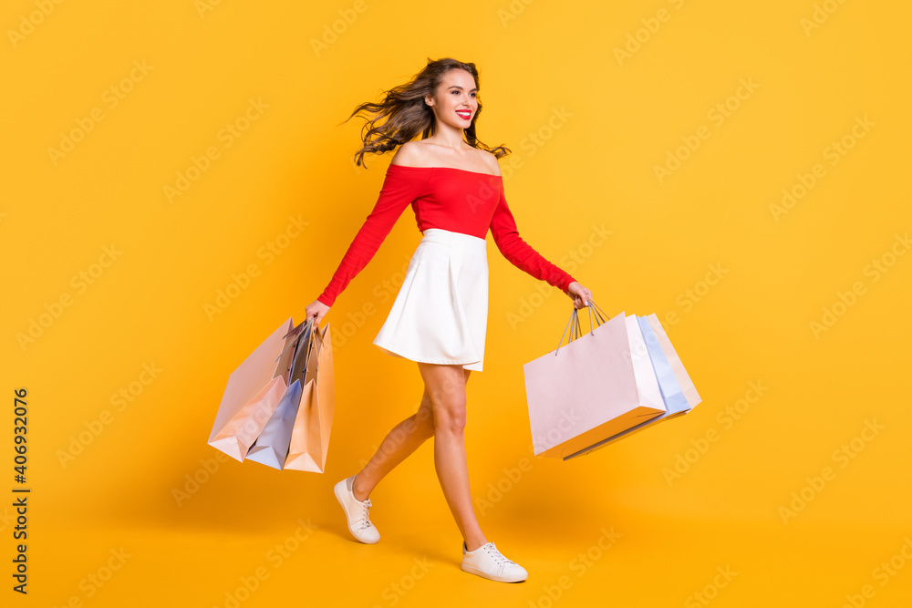 Full length body size photo girl smiling looking blank space stepping with packages from store isolated on bright yellow color background Full length body size photo girl smiling looking blank space stepping with packages from store isolated on bright yellow color background