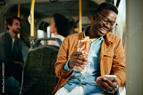 Foto Happy black man eating a sandwich and using smart phone while commuting by bus