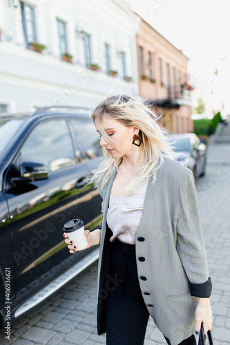 modern young blonde in the city is walking against the background of a car with a cup of coffee. © velimir
