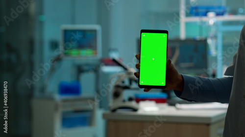 Close up of chemist's hand holding smartphone with green screen standing in biological laboratory while nurse bringing blood samples. Scientist using smartphone with mockup, chroma key display