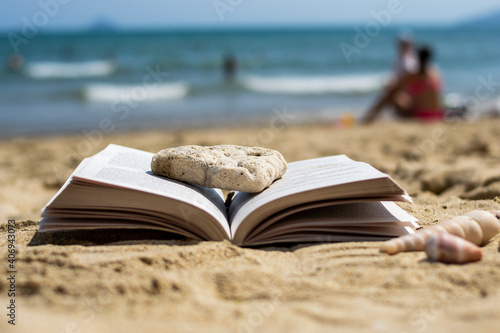 An open book crushed by a stone lies on the sand on the beach. Blurred background. Summer vacation on the beach