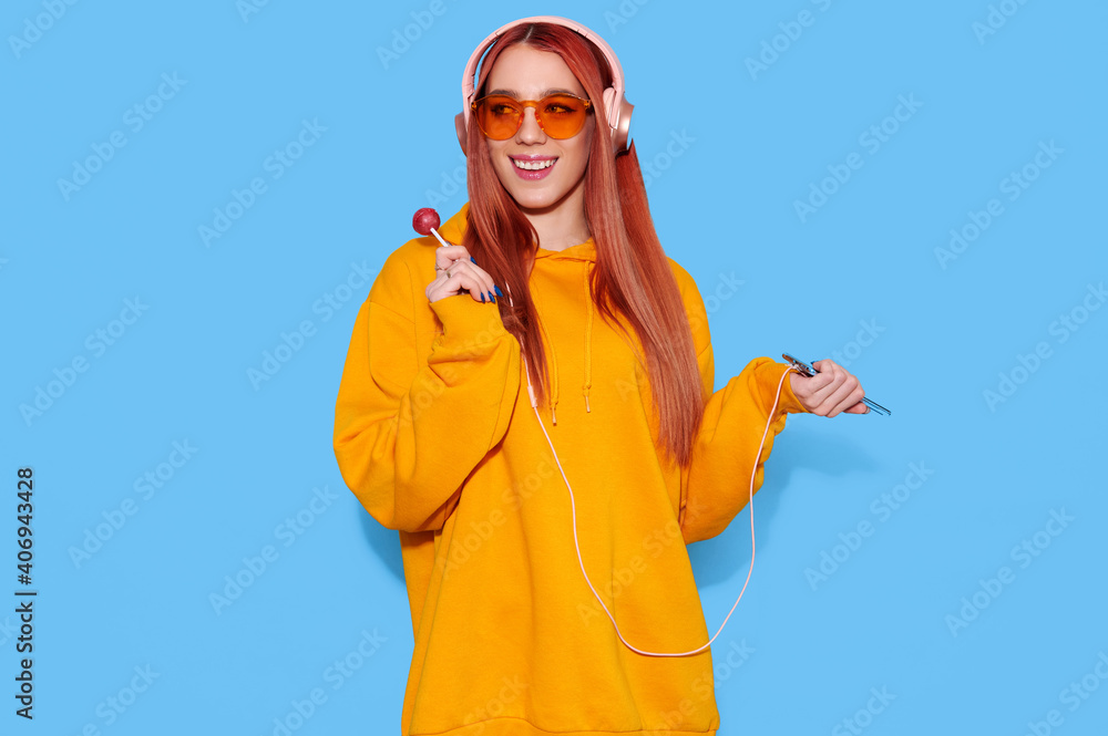 Hipster teenage girl wearing trendy oversize hoodie eats lollipop and listen to music in headphones on blue background Hipster teenage girl wearing trendy oversize hoodie eats lollipop and listen to music in headphones on blue background