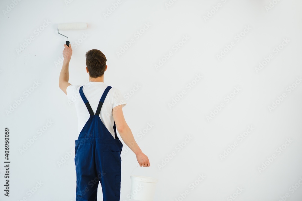 young man in blue overalls painting wall into white color with a roller young man in blue overalls painting wall into white color with a roller
