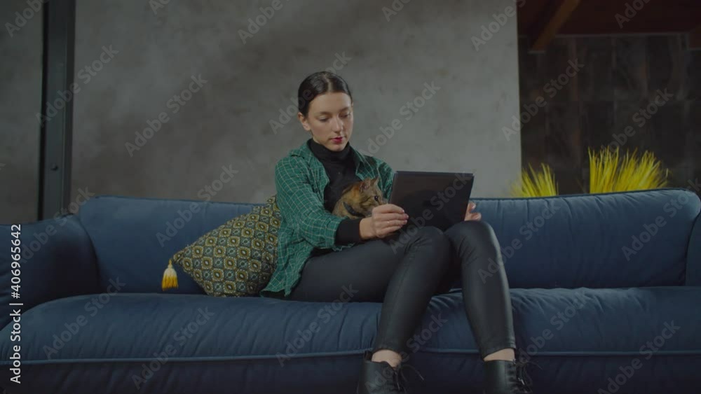 Positive happy charming female relaxing on couch, browsing online social media content on digital tablet and stroking cute shorthair tabby cat while spending leisure with pet in domestic room. Positive happy charming female relaxing on couch, browsing online social media content on digital tablet and stroking cute shorthair tabby cat while spending leisure with pet in domestic room.