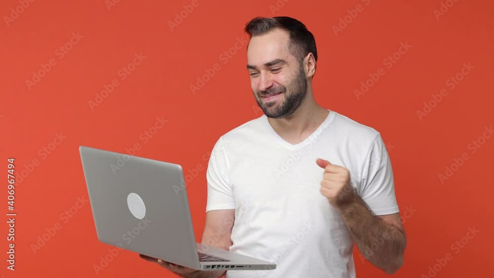 Funny bearded young man 20s in white casual t-shirt isolated on orange background studio. People lifestyle concept. Working on laptop pc computer making video call greeting with hand showing thumb up Funny bearded young man 20s in white casual t-shirt isolated on orange background studio. People lifestyle concept. Working on laptop pc computer making video call greeting with hand showing thumb up