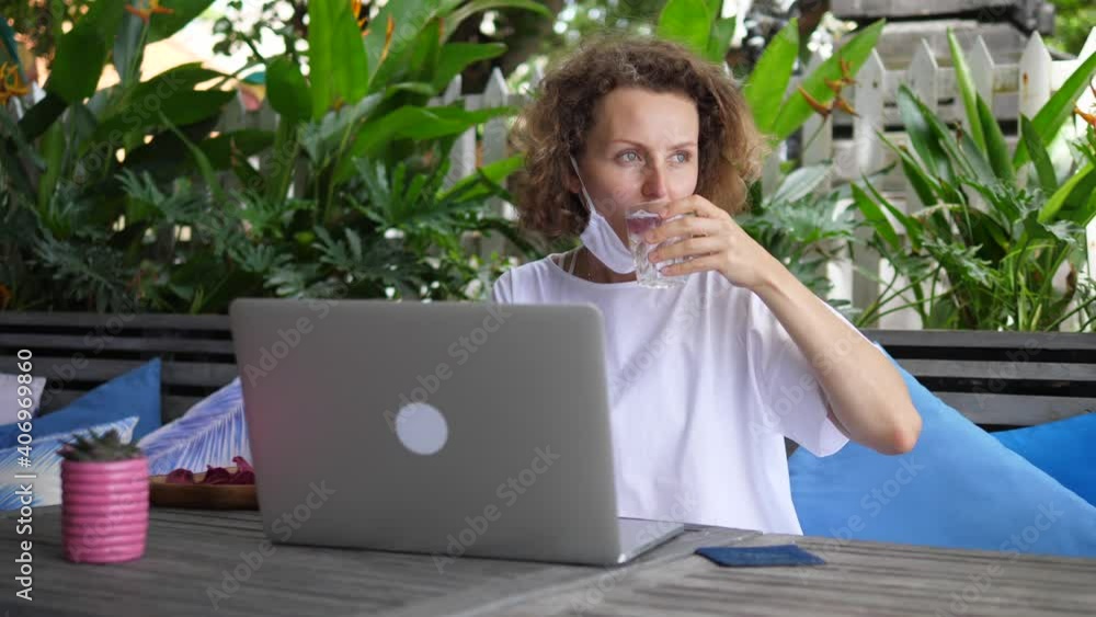 Caucasian female freelancer working on her laptop outdoors takes off face mask to drink water. Tropical interior Caucasian female freelancer working on her laptop outdoors takes off face mask to drink water. Tropical interior
