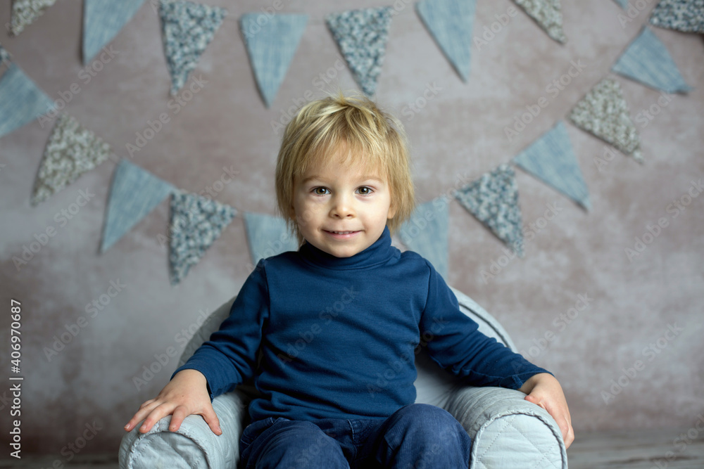 Naklejka premium Cute blond toddler child, boy, sitting on little baby armchair, reading a book and holding flowers