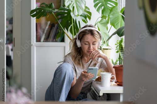 Woman listening to music, wear wireless white headphones, using mobile smart phone, chatting in social networks, sitting next to the window, houseplants on windowsill. Life at home. Time to relax.  photo