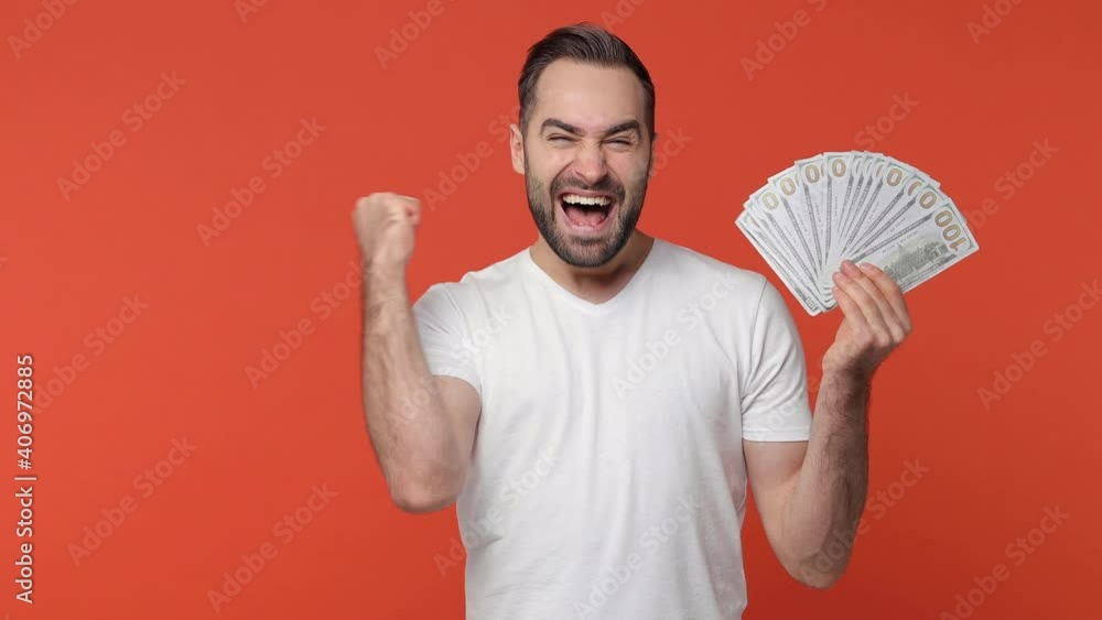 Happy overjoyed bearded young man 20s in white casual t-shirt isolated on orange background studio. People lifestyle concept. Hold fan cash money in dollar banknotes doing winner gesture celebrating Happy overjoyed bearded young man 20s in white casual t-shirt isolated on orange background studio. People lifestyle concept. Hold fan cash money in dollar banknotes doing winner gesture celebrating