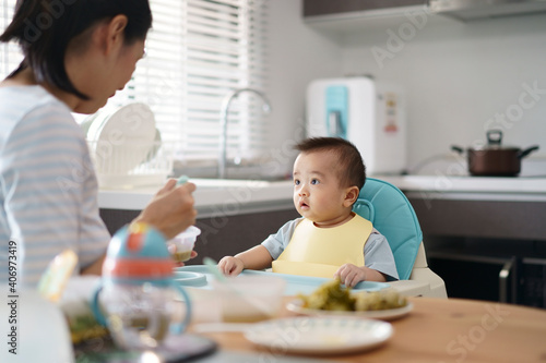 Asian young mother provide food and fruit puree for lunch to her baby seat on high chair in kitchen at home. The baby try to weaning and looking at his mom. A wife  take care her son.