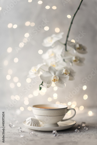 Cozy home spring breakfast still life. A porcelain cup of hot green tea with a marshmallow dessert on a gray background is decorated with a flowering branch of a white otchid. Spring concept. 