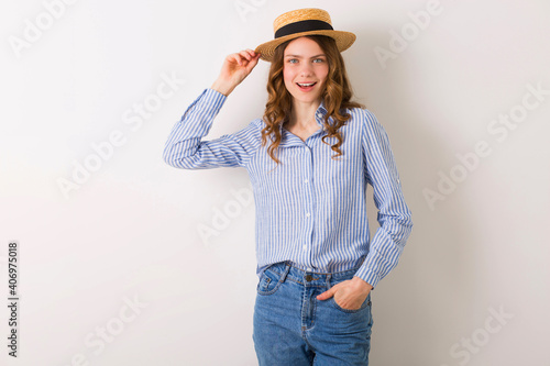 young beautiful stylish woman in summer style outfit posing on white wall wearing straw hat