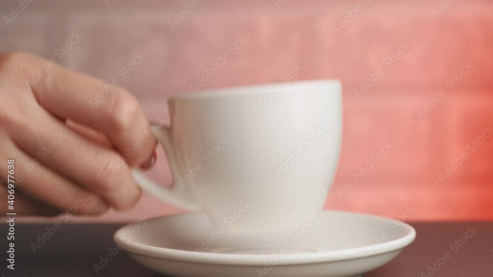 A woman's hand puts a white cup of coffee on the table. Red background and brick wall. Red light on the right. A woman's hand puts a white cup of coffee on the table. Red background and brick wall. Red light on the right.