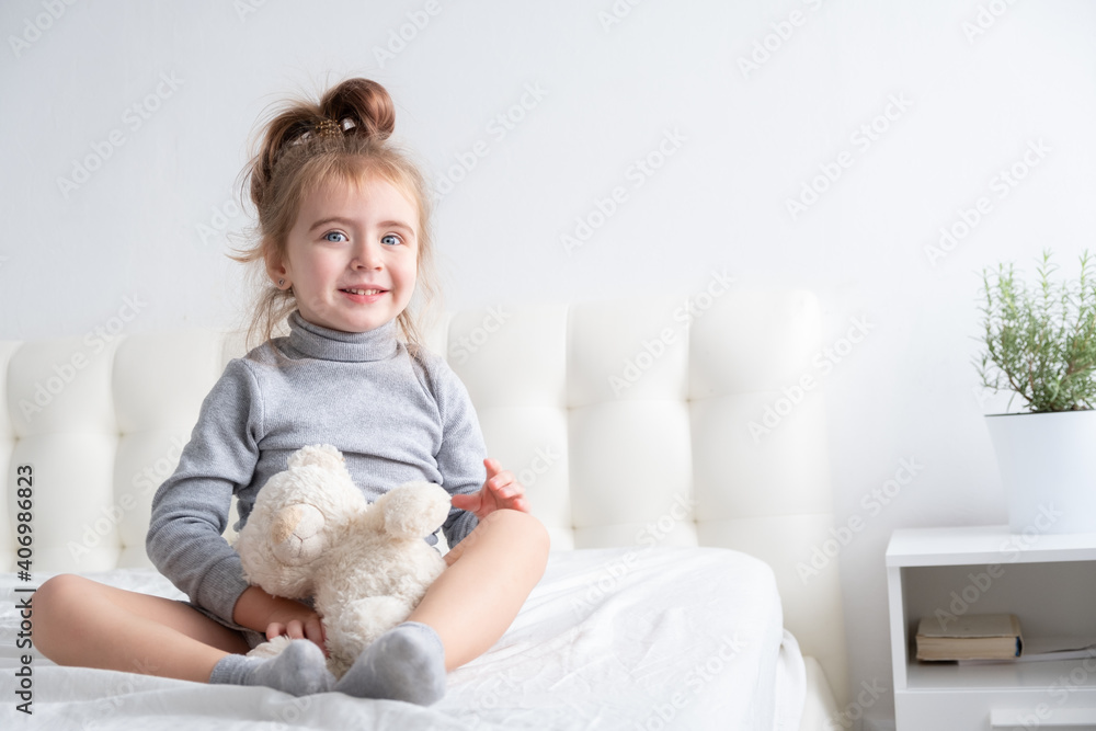 little girl in grey turtleneck playing with teddy bear on white bedding little girl in grey turtleneck playing with teddy bear on white bedding