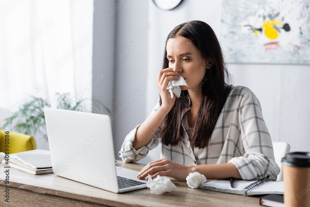allergic woman wiping nose with paper napkin while working at laptop on blurred foreground allergic woman wiping nose with paper napkin while working at laptop on blurred foreground