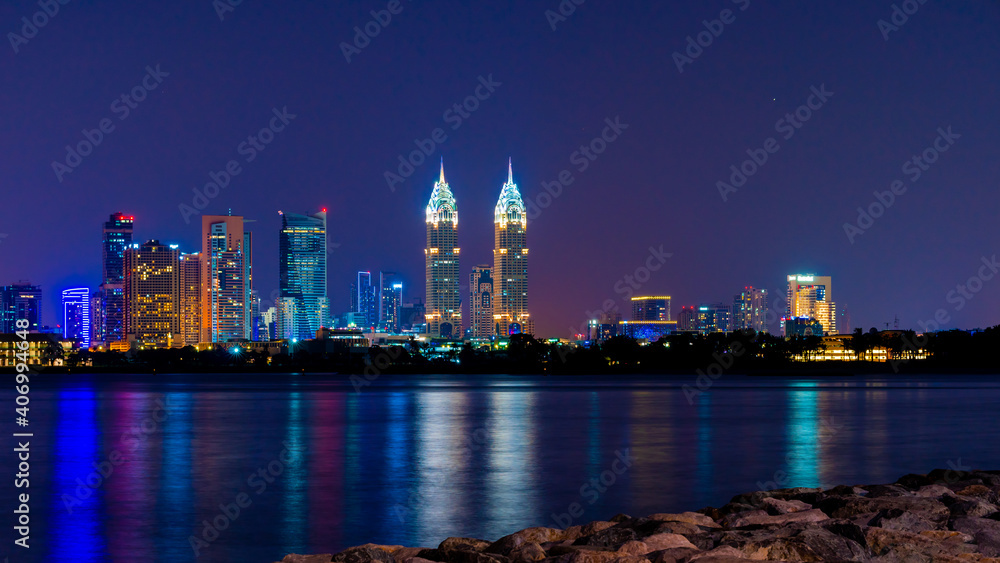 Night view to iconic skyscrapers panorama of Dubai. Amazing illumination of the Business Central Towers. Luxury skyscrapers represent modern Dubai. Colors reflect over water.