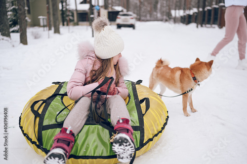 Little cute girl in pink warm outwear having fun with red shiba inu dog rides inflatable snow tube in snowy white cold winter outdoors. Family sport vacation activities.