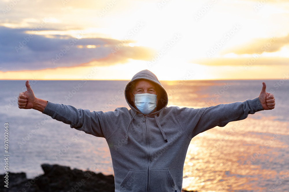 Front view of senior man with hood on head standing on top of cliff with ocean behind him. One people enjoying a winter day at sea with raised arms, wearing surgical mask due to coronavirus - obrazy, fototapety, plakaty 