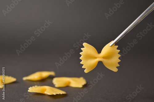 Piece of Uncooked italian farfale bowtie pasta in metal tweezers isolated on black background with four pieces of raw pasta on background
 photo