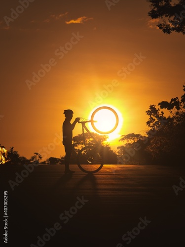 a man poses with his bicycle in a sunset, he lifts the bicycle and his tire is just ahead of the sunset.
