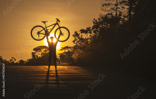 a man lifts his bicycle with his hands in the middle of a road with the sun behind it, an orange sun.