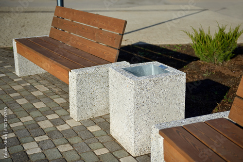 In the park there is a wooden bench with a back  with marble legs and a marble trash can.Outdoor bench made of wood and marble.White marble trash can with a metal surface inside. Wooden bench for rest