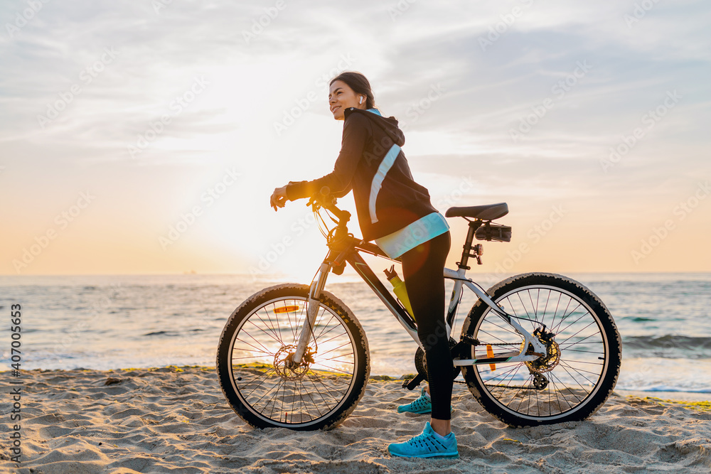 young attractive slim woman riding bicycle, sport on morning sunrise summer beach in sports fitness wear, active healthy lifestyle, smiling happy having fun young attractive slim woman riding bicycle, sport on morning sunrise summer beach in sports fitness wear, active healthy lifestyle, smiling happy having fun